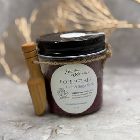 A jar of Raindrop to Remedy Rose Petals Herb & Sugar Scrub with a black lid, wrapped in twine with a small wooden spaoon